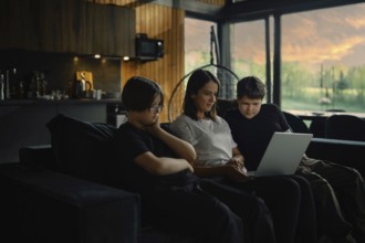 Young mother and her sons watch a series on a laptop sitting in the evening in a cozy living room