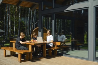 Two children and their mother are seated at a wooden table outside a modern building. They are