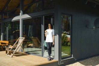 A woman stands on the deck of a stylish house, holding a drink while enjoying a sunny day. The home