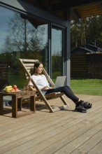 A woman sits comfortably on a deck chair, focused on her laptop. She is scrolling through social