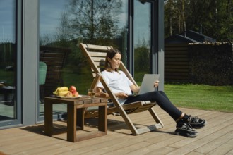 A woman sits comfortably in a deck chair on a wooden patio. She opened the laptop cover to check