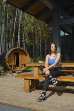 A woman dressed in casual sportswear sits comfortably on a wooden bench in front of a traditional