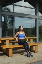 A woman sits on a wooden bench in front of a large glass cabin, basking in natural light.