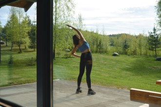 The view from inside the house at a woman is stretching her body on a wooden deck surrounded by
