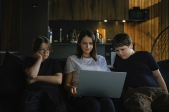 A woman and two children, are sitting closely on a sofa. They focus on a laptop screen in a warmly