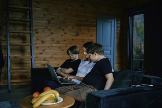 A family sits together on a comfortable couch in a wooden living room, focusing on a laptop. The