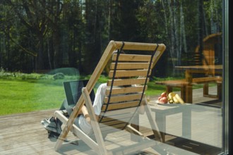 A person works on a laptop while reclining in a wooden lounge chair on a deck surrounded by trees.
