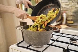 A person transfers sliced vegetables in a pot while adding more ingredients from a frying pan on a