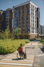 A man using a wheelchair navigates a crosswalk in a modern urban setting. The sun shines brightly,