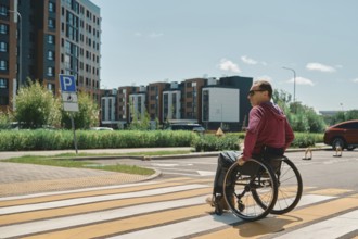 A man in a wheelchair is crossing a bright crosswalk on a sunny day. Surrounding him are modern