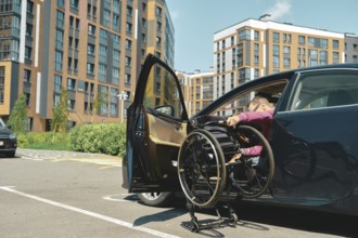 A person is maneuvering a wheelchair into a car parked in a contemporary urban area. A man unfolds