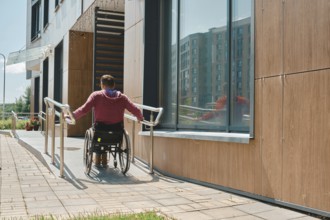 Rear view of a man in a wheelchair is maneuvering up a ramp beside a contemporary building. The