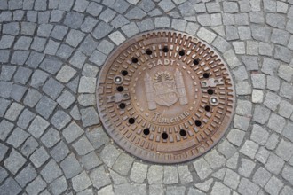 Round manhole cover with town coat of arms, inscription, cobblestones, floor, Ilmenau, Thuringian