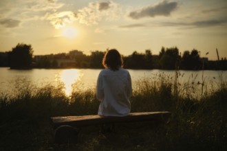 At the edge of a serene lake, a woman sits on a wooden bench surrounded by tall grass, soaking in