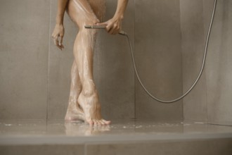 Water cascades over female legs in a sleek bathroom. The individual uses a handheld showerhead to