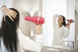 A woman is using a hair dryer to style her wet hair while standing in front of a large mirror. The