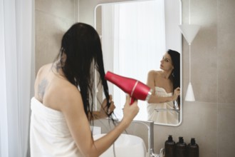 A woman stands in a bright, contemporary bathroom, holding a hair dryer and drying her wet hair.