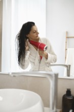 A woman in a white robe stands in a well-lit bathroom, carefully drying her wet hair with a bright