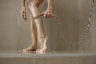 A woman stands in a sleek shower, washing soap off her legs while using a handheld showerhead.