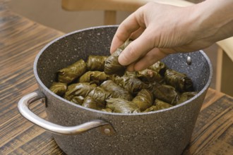A hand carefully adds stuffed grape leaves to a pot filled with dolma in a warm home kitchen. The