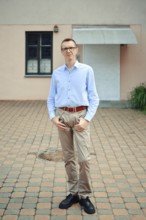 A man stands confidently outside a modern building, wearing a light blue shirt and beige pants. His