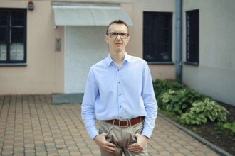 A man with glasses in a light blue shirt and beige trousers standing in front of a building. He has
