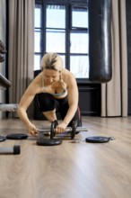 A focused woman is adjusting weights while kneeling on a gym floor. The space has large windows