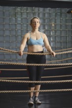 A woman in a blue sports bra and black leggings stands in a boxing ring with rope surrounds