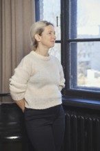 A woman with light hair is standing by a window, gazing thoughtfully outside. She is dressed in a