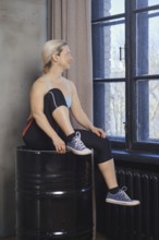 A woman sits on a barrel, pausing between exercises. She gazes out the large windows, showcasing