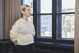 A woman stands next to a large window, lost in thought as she gazes outside. The sunlight filters