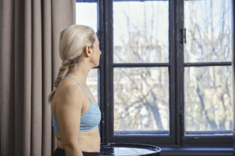 A woman with braided hair is standing near a large window, looking outside at the bright day. She