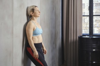 A woman leans against the wall in a modern studio, focusing on her breath as sunlight filters