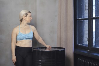 A woman wearing a blue sports bra and black leggings stands next to a black barrel, gazing