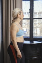 A woman stands near a window, wearing a sports bra and leggings, looking thoughtfully outside. The