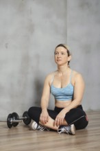A fit woman is seated cross-legged on the gym floor, looking determined and focused