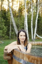 A woman enjoys a tranquil moment in a wooden hot tub, framed by tall trees and lush greenery during