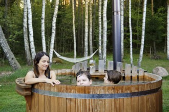 A woman and two children share a joyful moment in a wooden hot tub surrounded by trees. The