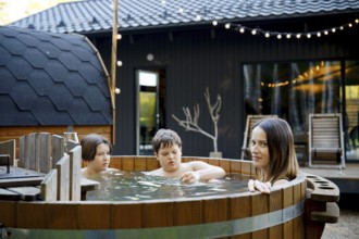 Mother and two children relax in a wooden hot tub in a backyard. The warm water scented with fir