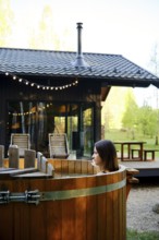 A woman enjoys relaxing in a wooden hot tub in the backyard of a tiny house. The sun illuminates