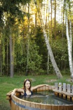 A woman relaxes in a wooden hot tub nestled in a forested area. The warm sunlight filters through