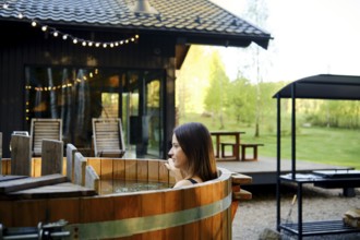 A woman sits in a rustic wooden hot tub surrounded by greenery at a countryside retreat. The