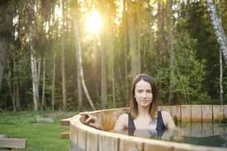 A woman enjoys a soothing soak in a wooden hot tub amidst tall trees during a serene sunset. The