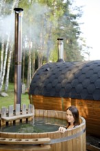 A woman enjoys a peaceful moment in an outdoor wooden hot tub surrounded by tall trees. Steam rises