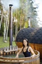 A woman enjoys a peaceful moment in a wooden hot tub, soaking in warm water. The nearby sauna sends