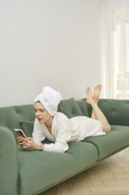 A woman in a white bathrobe and towel is lounging on a green couch, scrolling on her smartphone.