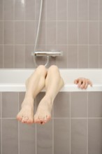 A pair of bare legs rests over the edge of a bathtub, with feet comfortably hanging. A hand is seen