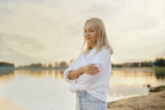 A young woman stands by the calm lakeside at sunset, wrapped in a cozy white shirt and denim jeans,