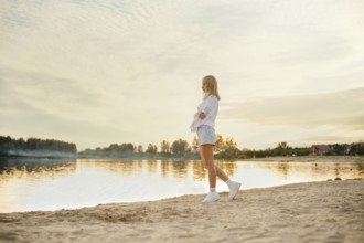 A young woman explores a sandy beach as the sun sets over a calm lake. The scenery features