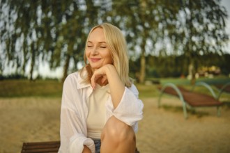 A pleased woman sits comfortably on a bench by the lake, smiling softly as the sun sets